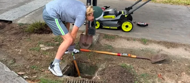 A student digging out a clogged storm drain beside a sidewalk, with shovels and a lawn mower nearby